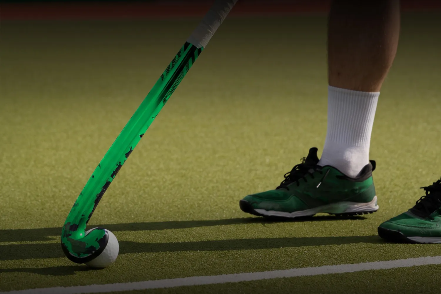 Palo de hockey verdinegro y zapatillas verdes sobre la cancha de césped sintético